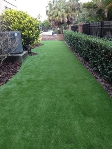 A clean, green artificial grass pathway between bushes and a wooden fence.