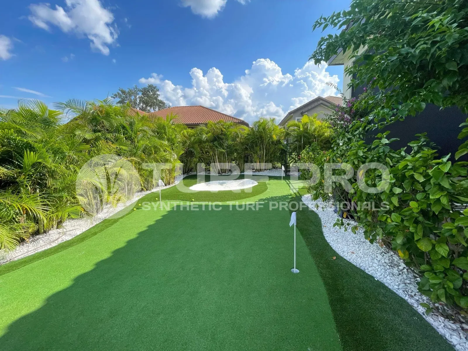 A backyard putting green with a flagstick surrounded by greenery and a stone wall.